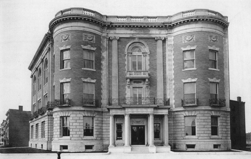 A black and white photo of the large, sturdy Massachusetts Historical Society building. A few small buildings are visible in the background.