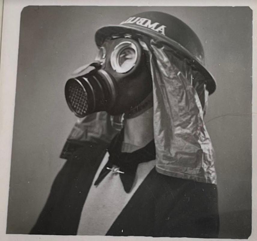 Black and white photograph of a white woman in a 1940s style gas mask. She is wearing a black jacket, light sweater, and a black tie. 