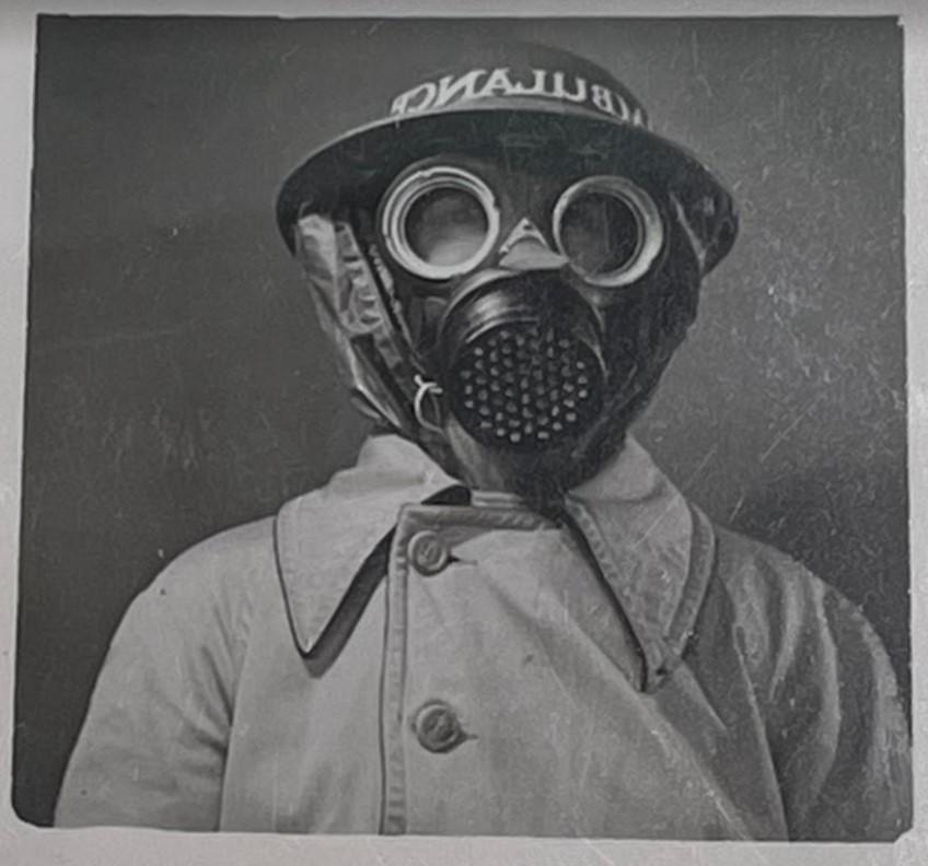 A black and white photograph of a woman wearing a 1940s style gas mask. She is wearing a light-colored buttoned-up trench coat. 