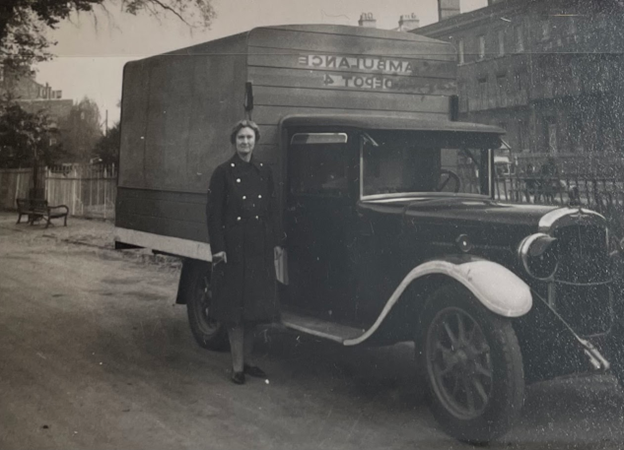 Black and white photograph of a middle-aged white woman standing next to a 1940's truck with "Ambulance, Depot 4," written on the wooden extension over the bed of the truck. She is wearing a long dark coat. 