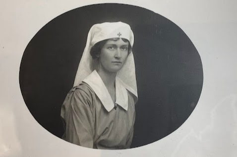 Color photograph of a black and white photograph in an oval shape. The image is of a young white woman with dark hair under a nurse's white habit with a medical cross at her temple. She wears a smock style dress with a wide white collar. The background is very dark. 