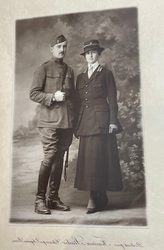 Color photograph of a black and white photograph of a man and a woman standing together in front of a painted backdrop of trees and a field. She stands to the left in a military red-cross uniform, a long skirt, flared jacket, white shirt and light tie and a hat with a brim and the Red Cross cross on it. To the right the man is in a WWI military uniform with high leather books, flared pants at the hips - tight at the knee, a long military jacket with leather belt and shoulder strap and hat. 