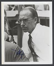 small black and white image of a white man with thinning hair wearing a white shirt with a dark tie. He is wearing large glasses on his face and is frowning